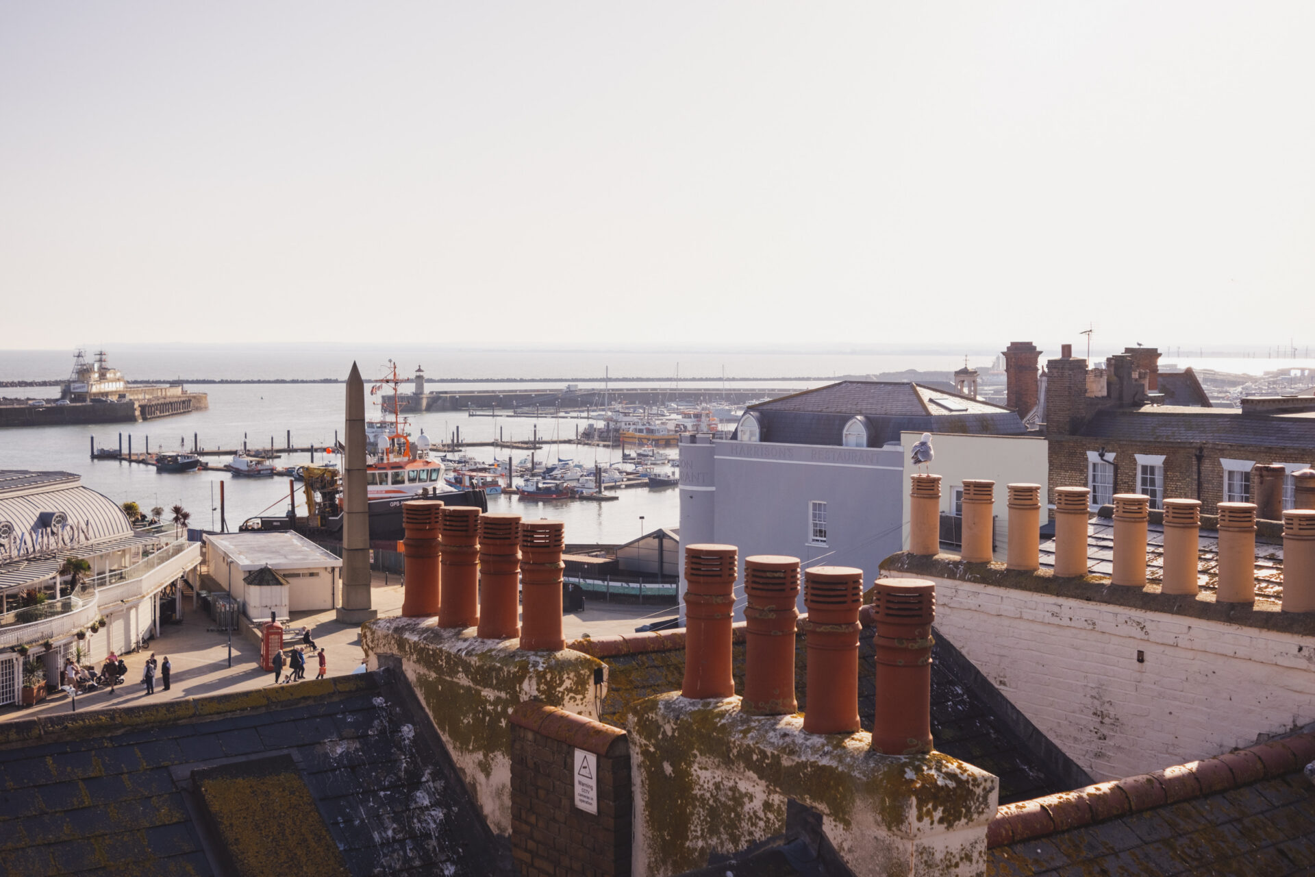 A sunny, high-angle view looking over mossy rooftops and terracotta chimneys towards Ramsgate's waterfront. Below, pedestrians walk near the Ramsgate Obelisk, a red telephone box, and the Royal Victoria Pavilion. In the background, the Royal Harbour is filled with moored boats and a large workboat, with the sea stretching out to a hazy horizon.