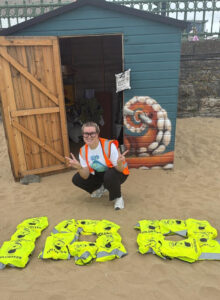 Rise Up Clean Up - A smiling volunteer in front of a beach hut with hi vis vest laid out on the beach, during RUCU’s 100th clean.