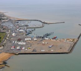 High-angle aerial shot of the Port of Ramsgate, featuring a town on white cliffs, a marina full of boats, and a commercial port area with trucks enclosed by a sea wall.