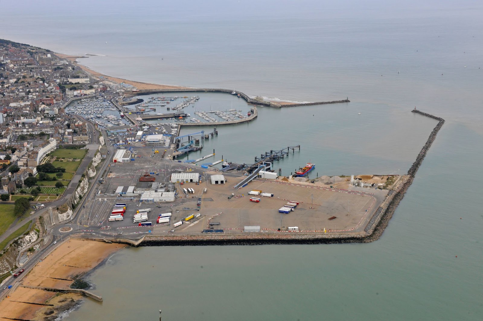 High-angle aerial shot of the Port of Ramsgate, featuring a town on white cliffs, a marina full of boats, and a commercial port area with trucks enclosed by a sea wall.