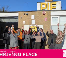 A group of ten people cheer with arms raised outside a brick building featuring a large yellow PIE sign.