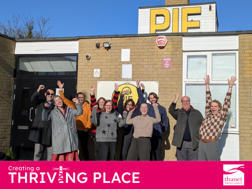 A group of ten people cheer with arms raised outside a brick building featuring a large yellow PIE sign.