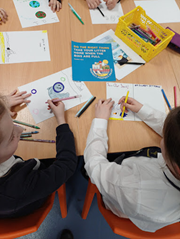 An overhead view of students seated at a desk, drawing and coloring on paper. In the center of the table is a blue leaflet that reads 'DO THE RIGHT THING TAKE YOUR LITTER HOME WHEN THE BINS ARE FULL'.