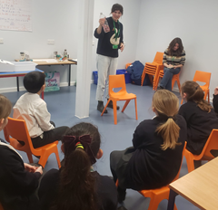 An instructor stands in front of a group of seated students, holding up a plastic water bottle to show them. She is wearing a dark top with a swan design and a green lanyard. Another adult sits in the background, and the students are sitting on bright orange chairs in a classroom setting.