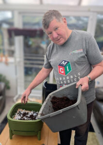 An man standing in a bright sunroom, holding a grey tub filled with dark compost or soil. He is looking toward the camera while adding the compost into a green, multi-tiered indoor composting bin. He is wearing a grey t-shirt.