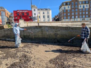 Two people using litter pickers and clear plastic bags to collect rubbish on a sandy, seaweed-covered beach on a sunny day. They are standing in front of a stone wall, behind which are several multi-story buildings, including a prominent bright red building with 'The Old Kent Market' written across the front.