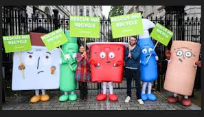 A group of two people and five large mascots standing outdoors on a paved area in front of a black metal gate. The mascots are dressed as recyclable items with sad faces, including a white carton, a green bottle, a red tub, a blue spray bottle, and a cardboard tube. Several of the mascots and the people are holding bright green signs that read 'RESCUE ME! RECYCLE'.
