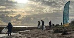 A silhouetted group of people participating in a beach clean-up under a dramatic, cloudy sky with the sun low on the horizon. The volunteers are walking along the shoreline holding white collection bags, and a tall, teal promotional flag stands on the grass to the right.