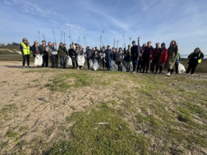 A large group of volunteers standing together outdoors on a grassy shoreline, smiling and posing for a photo on a sunny day. They are holding up litter-picking sticks and white collection bags, and a few people on the edges are wearing yellow high-visibility vests.