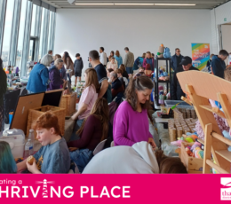 A bustling indoor market filled with people browsing stalls of handmade crafts, crochet items, and baked goods.