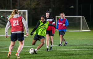 An evening women's football practice on an artificial grass pitch under floodlights. In the center, a player wearing a lime green training bib over a black shirt dribbles the football forward. She is closely followed by a defender in a black and red hooded jacket, while another player wearing a red training bib with the number 21 watches from the foreground.