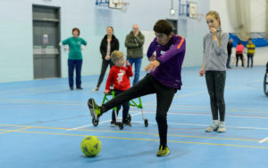 An indoor sports hall with a blue floor where a young man in a purple long-sleeve athletic shirt is kicking a yellow football. Behind him, a young boy in a red hoodie stands using a green wheeled walker and watches the ball. A teenage girl and three adults stand on the sidelines in the background observing the session.