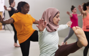 A diverse group of women participating in an indoor yoga or fitness class, practicing a balancing pose with their arms outstretched. In the foreground, a woman wearing a dusty pink hijab and a light green long-sleeve shirt focuses on holding her leg up. Behind her, another woman in a bright orange t-shirt performs the same pose, with other participants visible in the background.
