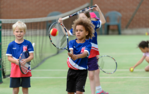 An outdoor green tennis court where young children are practicing next to a net. In the center, a young boy with curly hair focuses intensely as he swings a tennis racket at a red and yellow tennis ball mid-air. To his left, a smiling blonde boy stands holding his racket. Both wear matching blue and red "Tennis for Kids" shirts. Other children are visible practicing in the blurred background.