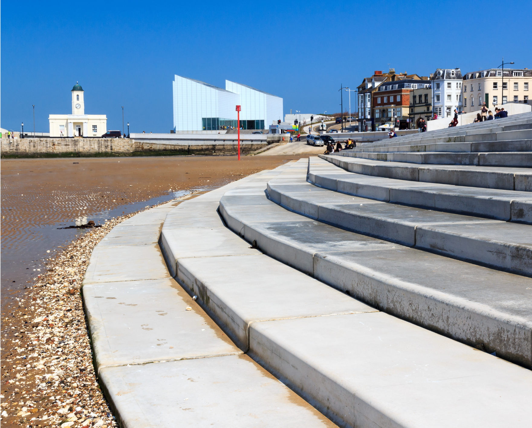 Wide, curving concrete sea steps lead down to a sandy beach in Margate, with modern and historic buildings behind.