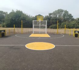 Front view of a outdoor tarmac sports court featuring a basketball hoop, football goal, and yellow painted markings.
