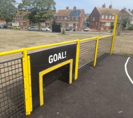 Close-up of a small mini-football goal panel built into yellow fence surrounding an outdoor sports court.