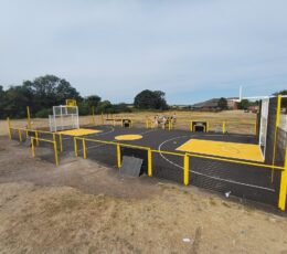 Wide angle shot of a black tarmac multi-use games area with yellow fencing and sports court markings.