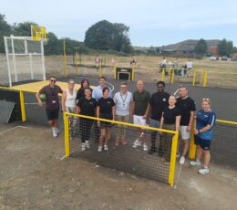 A group of eleven people pose together for a photo behind a low yellow fence at a sports court.