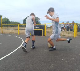 Two boys wearing grey football kits play football together on an outdoor court surrounded by a yellow fence.