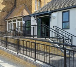 Exterior view of a newly built white modern extension connected to a period yellow-brick building with scaffolding on the left. A gently sloping accessibility ramp with black metal railings runs alongside a small flight of steps, both with dark railings, leading up to an open sliding glass entrance door under a dark canopy.