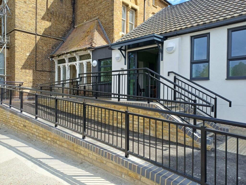 Exterior view of a newly built white modern extension connected to a period yellow-brick building. A sloping accessibility ramp with black metal railings runs alongside a small flight of steps, both with dark railings, leading up to an open sliding glass entrance door under a dark canopy.