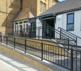 Exterior view of a newly built white modern extension connected to a period yellow-brick building. A sloping accessibility ramp with black metal railings runs alongside a small flight of steps, both with dark railings, leading up to an open sliding glass entrance door under a dark canopy.