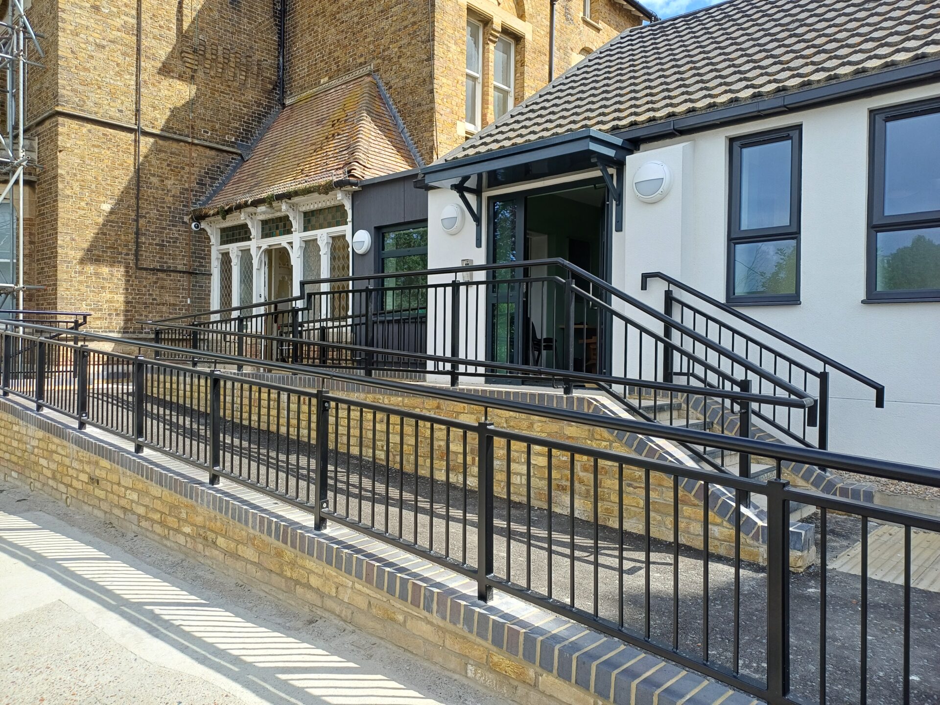 Exterior view of a newly built white modern extension connected to a period yellow-brick building. A sloping accessibility ramp with black metal railings runs alongside a small flight of steps, both with dark railings, leading up to an open sliding glass entrance door under a dark canopy.