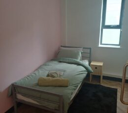 A single bed with sage green bedding and a light-coloured towel sits against a pink wall. To the side are a light-wood bedside table and a bright window.