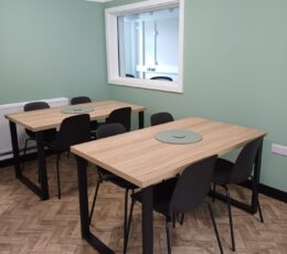 A bright room with sage green walls, wooden flooring, and two light wood tables surrounded by black chairs. Each table has a circular grey mat in the centre.