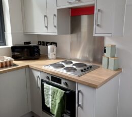 A clean, modern kitchen area with light grey cabinets, wooden worktops, a built-in oven with an electric hob, and green tea, coffee, and sugar canisters.