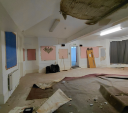 Interior room stripped for restoration work with damaged ceiling plaster, stripped wall panels, and floor protection.