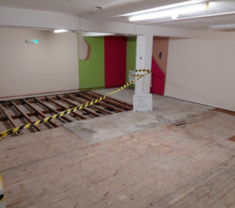 Interior view of building renovation showing exposed floor joists cordoned off with safety tape and a "keep out hole in floor" sign.