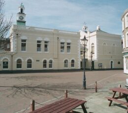 Margate Museum's cream-coloured historic building features a clock tower overlooking a paved square, with wooden picnic tables and a traditional lamppost in the foreground.