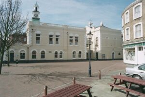 Margate Museum's cream-coloured historic building features a clock tower overlooking a paved square, with wooden picnic tables and a traditional lamppost in the foreground.