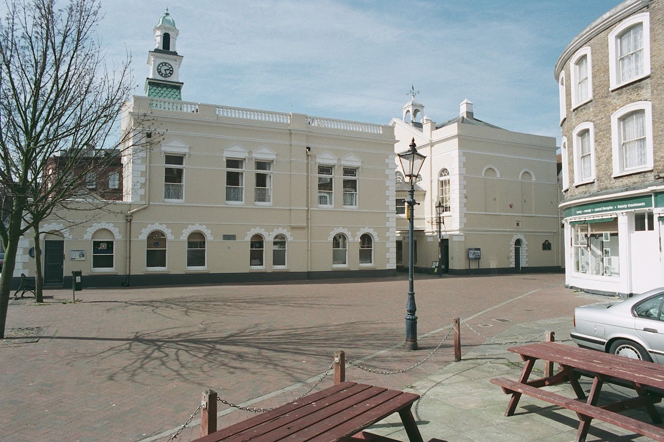 Margate Museum's cream-coloured historic building features a clock tower overlooking a paved square, with wooden picnic tables and a traditional lamppost in the foreground.